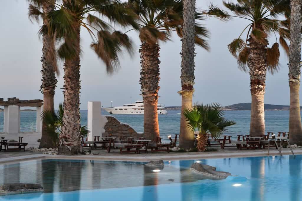 A luxurious seaside resort with a pool surrounded by tall palm trees at dusk. Wooden lounge chairs and picnic tables are arranged along the waterfront with evening light