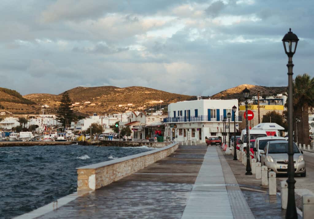 A coastal town scene in Parikia, Paros, featuring white buildings with blue shutters, cars parked along the street, and a view of the sea and mountains under a cloudy sky.