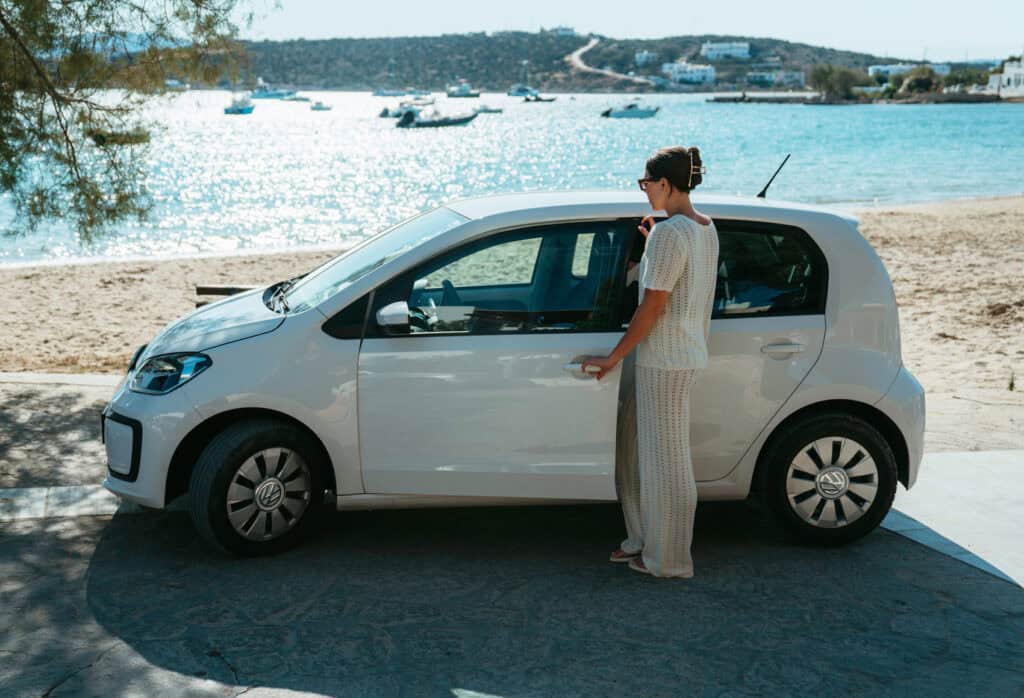 Sarah opening the door of a white rental car parked by the beach in Paros, Greece