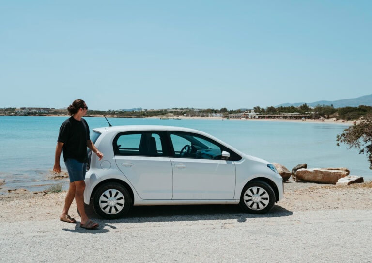 Caleb standing next to a white rental car by the sea in Paros, Greece — highlighting the ease and freedom of renting a car in Paros to explore the island’s beaches.