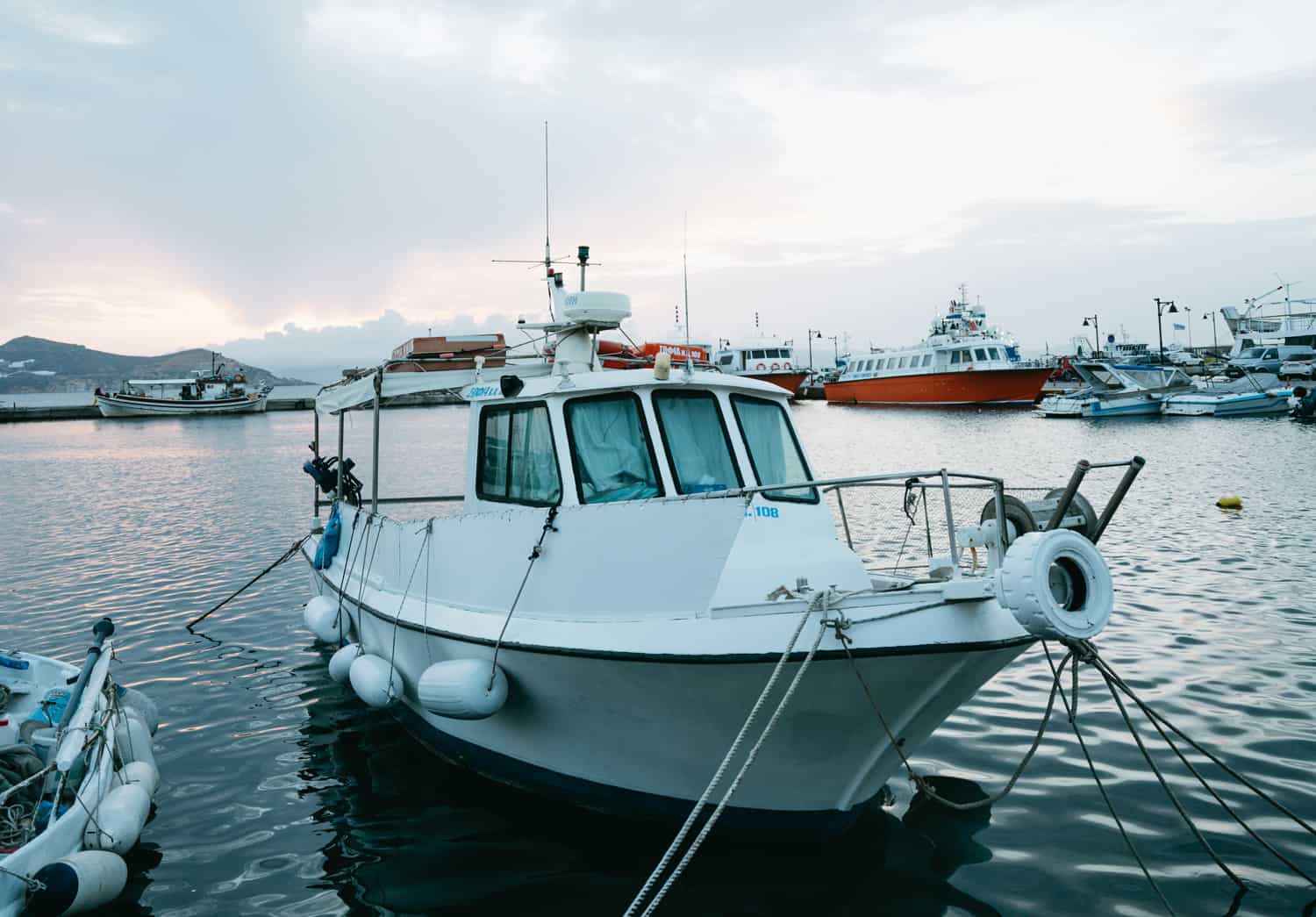 A small white fishing boat docked in a harbor, with other boats and ferries in the background during a calm evening, under a cloudy sky with the sunset in the distance.