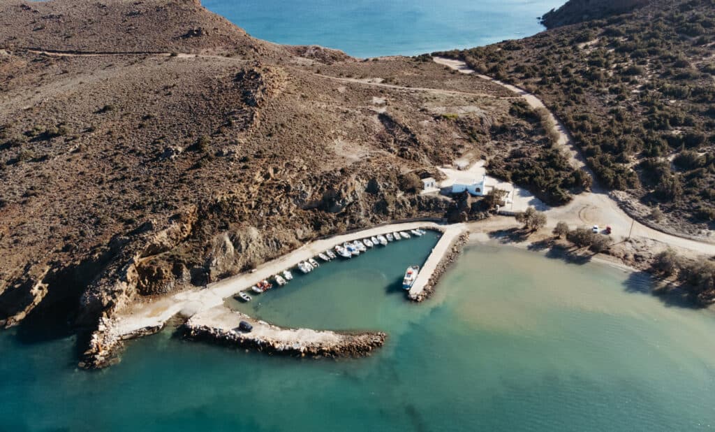 a drone view of Moles beach in Paros, Greece. there are mountains surrounding the shore and boats docked along the pier