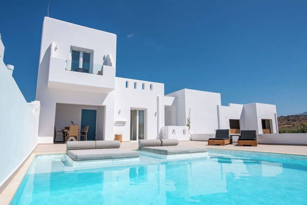 Depis Edem Luxury Villa's pristine white exterior, reflecting the iconic Cycladic architecture of the Greek islands. A stunning private pool with built-in sun loungers sits in the foreground, surrounded by minimalist outdoor furniture and a bright blue sky. The villa features multiple levels, a balcony with glass railings, and a shaded patio area.