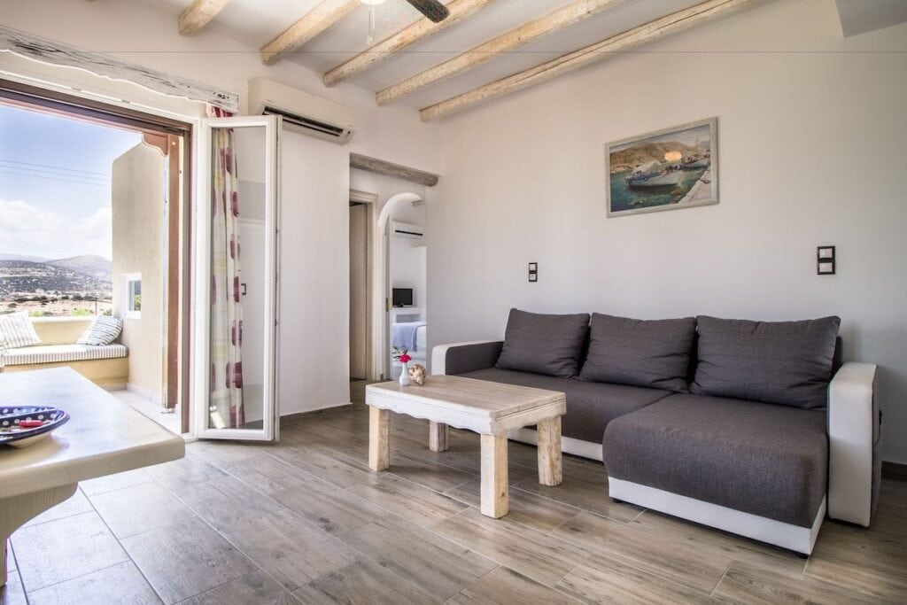 A stylish Mediterranean-style living room with rustic wooden beams on the ceiling, a gray sectional sofa, and a wooden coffee table. The space opens to a sunlit balcony with built-in seating