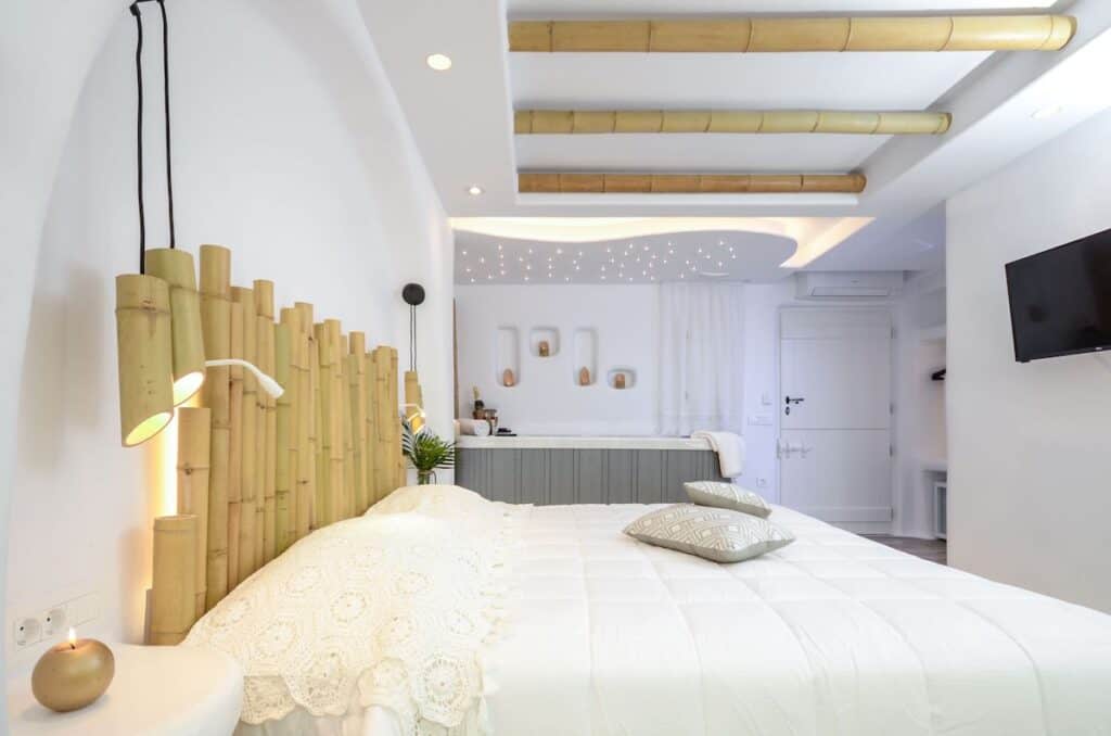 A bedroom at Naxos Island Escape Suites with a natural and minimalist aesthetic. The room features a bed with a white crocheted throw blanket and plush pillows, set against a bamboo headboard with matching pendant lights. The ceiling incorporates bamboo beams, and in the background, a small kitchenette