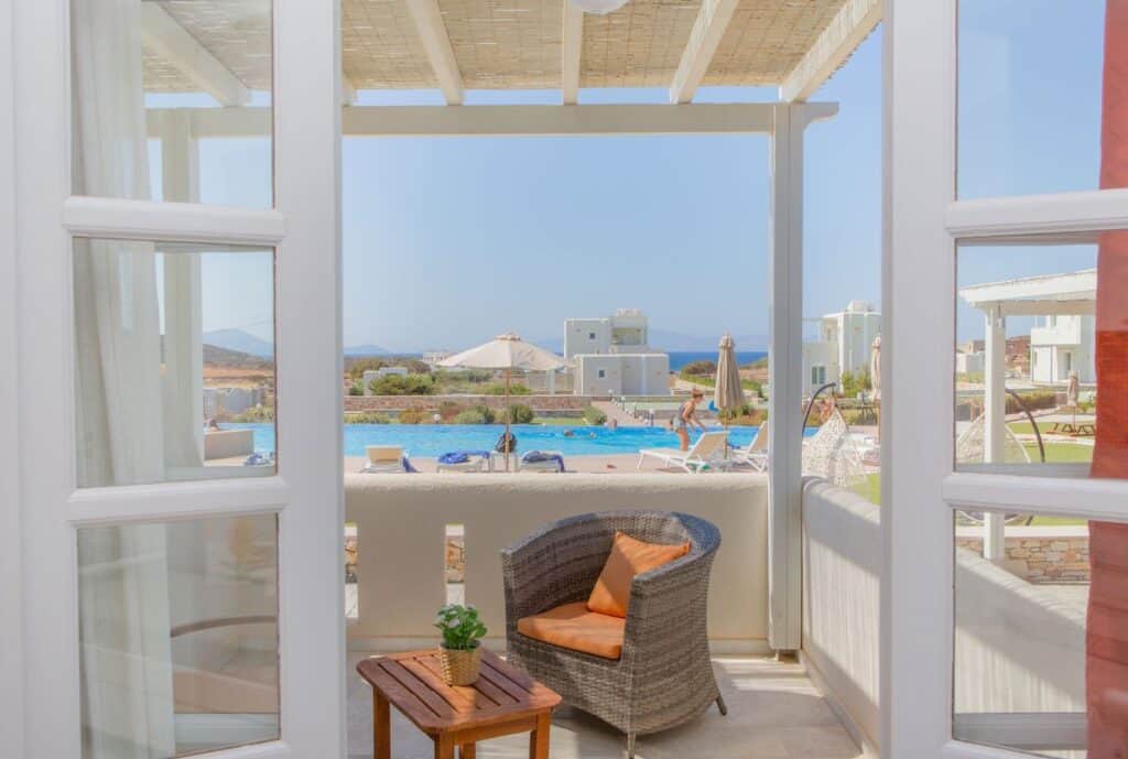 A bright and airy resort view seen from a shaded patio with a wicker chair and wooden table. The scene overlooks a pool with lounge chairs and umbrellas, surrounded by modern white villas against a backdrop of rolling hills and a clear blue sky. 