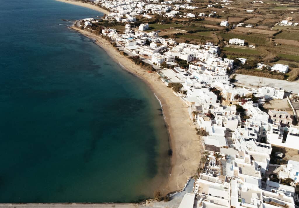 Aerial view of Agia Anna in Naxos, Greece, showing a curving sandy beach adjacent to stunning ocean. The coastline is lined with white buildings.