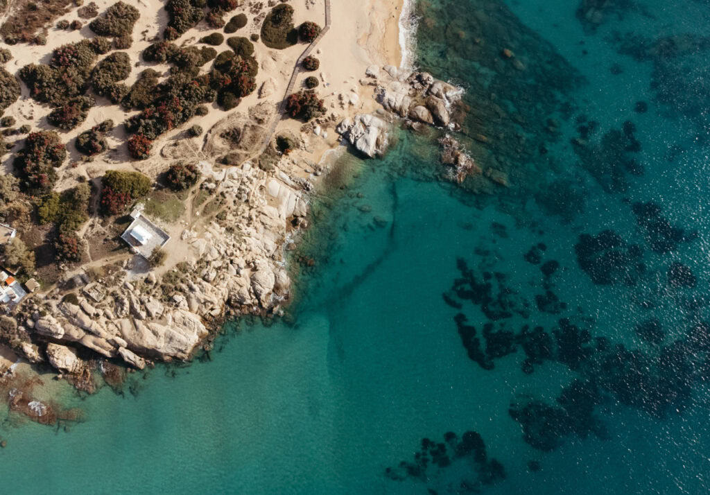 Aerial view of a rugged coastal area at Agia Anna, Naxos, showing a mix of sandy beaches and rocky outcrops along the ocean. The landscape is interspersed with lush greenery and pathways.