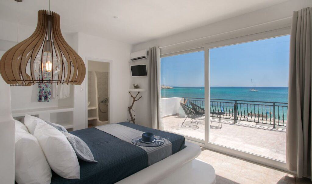 Manios Suites' beachfront bedroom with floor-to-ceiling glass doors opening to a private balcony overlooking the turquoise sea. The outdoor balcony has black chairs, offering a perfect spot to enjoy the ocean view with a sailing boat in the distance.
