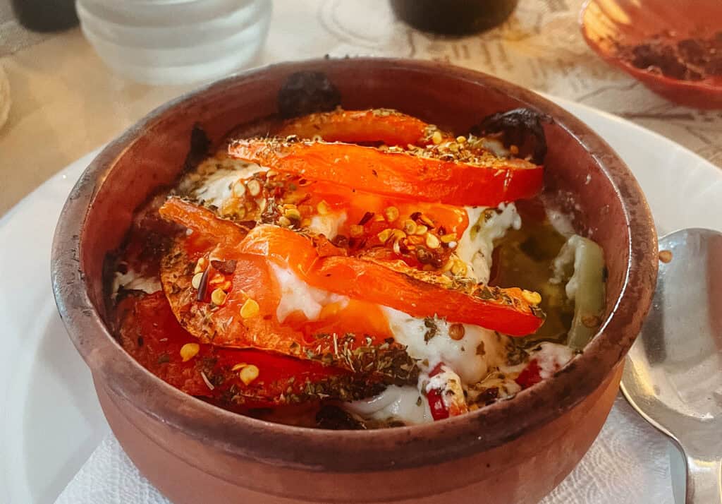 Close-up of a traditional Greek dish, baked feta cheese, served in a clay pot. The dish is topped with sliced tomatoes, herbs, and chili flakes, with the melted feta visible underneath.