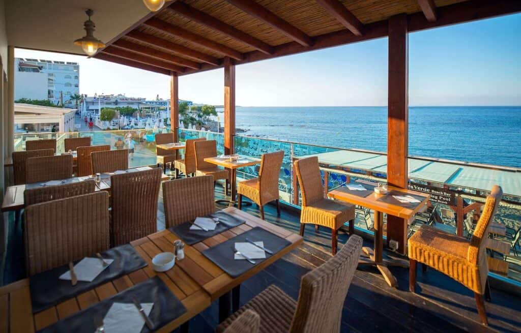 The open-air restaurant terrace at Golden Beach Hotel, overlooking the turquoise sea and a lively beach filled with sun loungers and umbrellas. The terrace features wooden tables set with black placemats, napkins, and silverware, surrounded by wicker chairs under a rustic wooden roof.