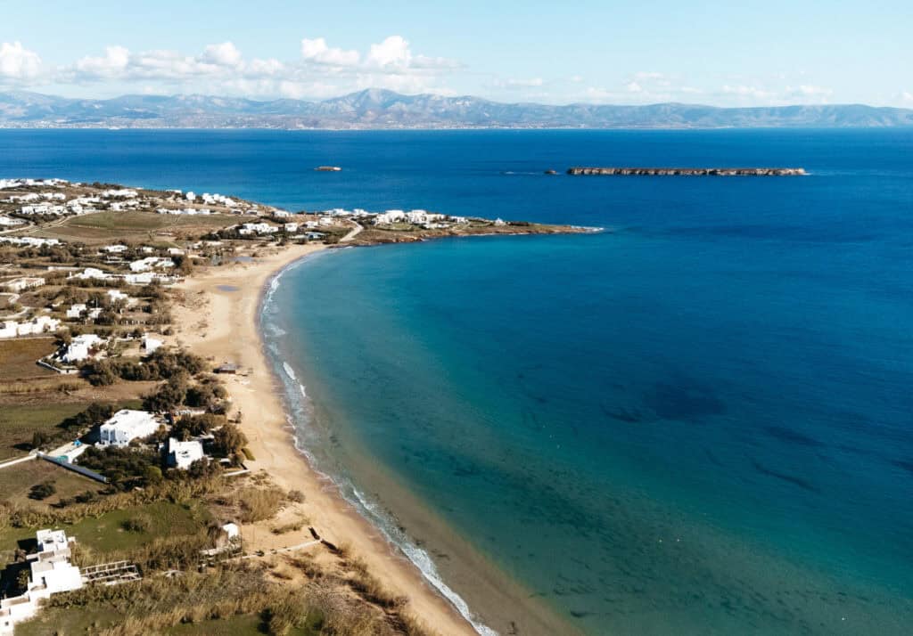 Aerial view of Golden Beach in Paros, Greece, showcasing a long, curved sandy shoreline with clear blue waters. The surrounding area includes white buildings and green fields, with a few small islands visible in the distance.
