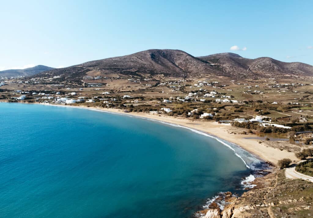 Aerial view of Golden Beach in Paros, Greece, featuring a wide sandy shoreline with clear blue waters. The surrounding area includes white buildings and green fields, set against a backdrop of rolling hills.