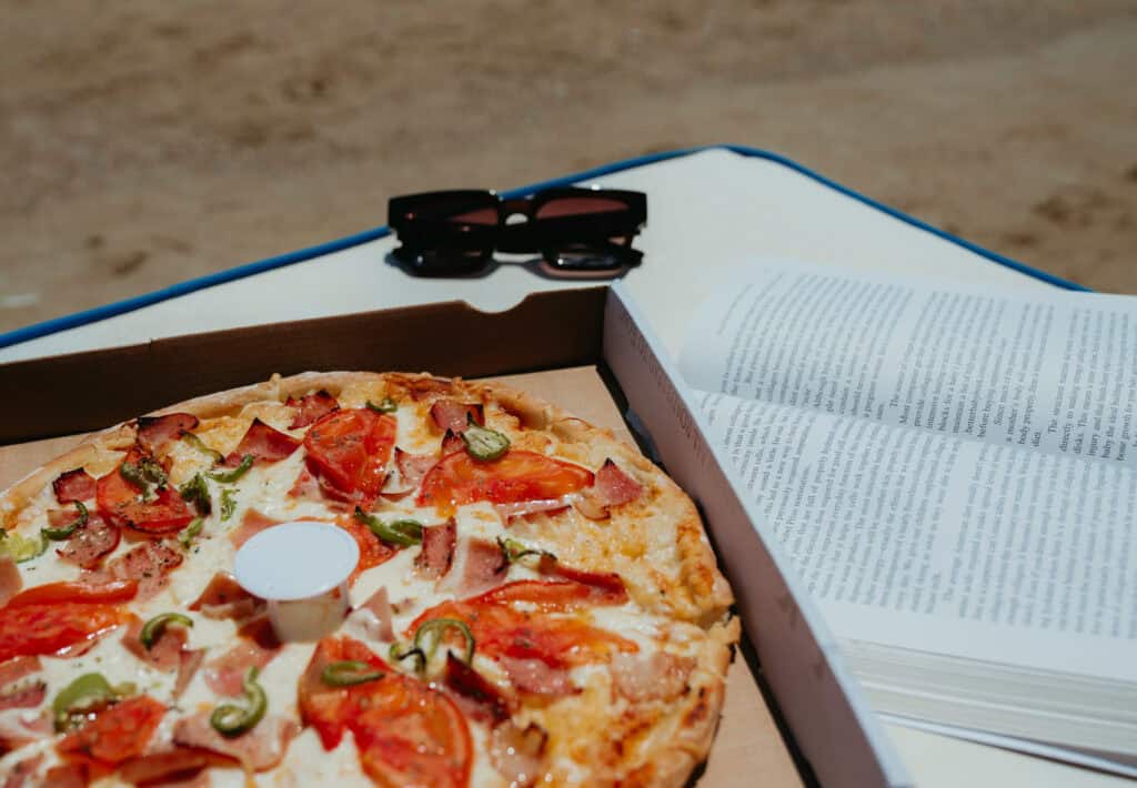 Pizza, sunglasses, and an open book on a beach lounger at Golden Beach Paros, perfect for a relaxing lunch by the sea.