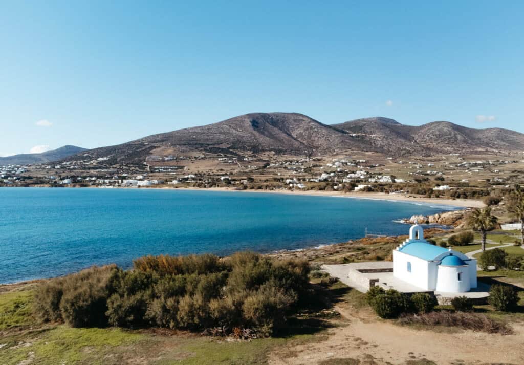 Aerial view of Golden Beach in Paros, Greece, featuring a small white chapel with a blue dome near the shoreline. The beach has clear blue waters, and the background shows rolling hills with scattered white buildings and green fields.