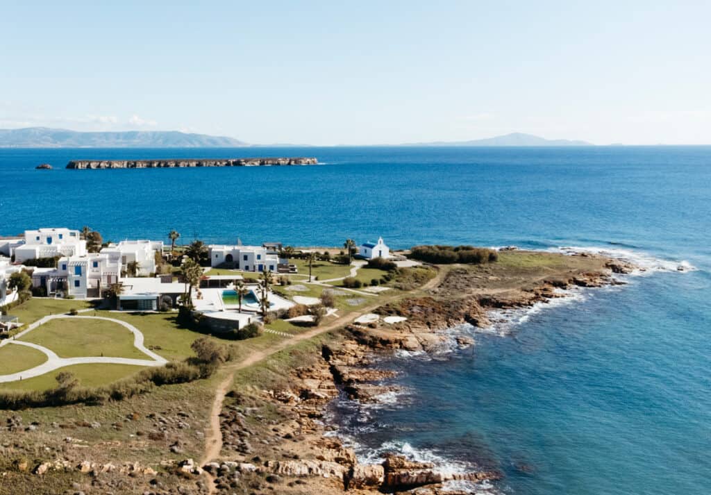 Aerial view of a coastal area in Paros, Greece, featuring white buildings and green lawns near a rocky shoreline. A small white chapel with a blue dome is situated near the water. The background includes a clear blue sea with a distant island and mountains under a bright, clear sky.