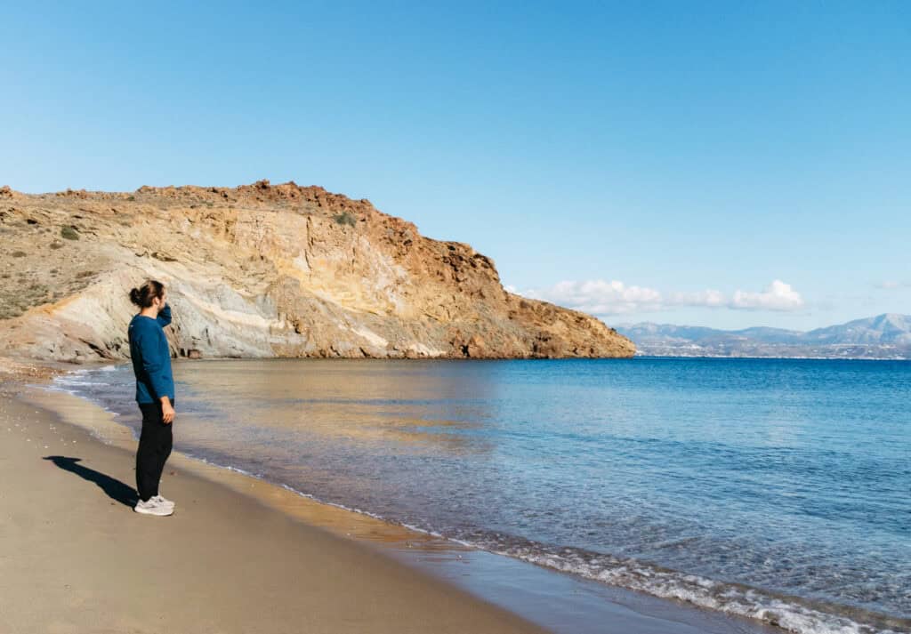 Caleb stands on the sandy shore of Kalogeros Beach in Paros, Greece, gazing at the sea. The beach is bordered by rocky cliffs with shades of brown and gray, and the clear blue water stretches out to the horizon.