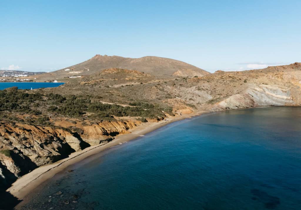 Aerial view of Kalogeros Beach in Paros, Greece, featuring a secluded, curved sandy shoreline with clear blue water. The beach is bordered by tall cliffs and hilly terrain, with a few white buildings visible in the distance.
