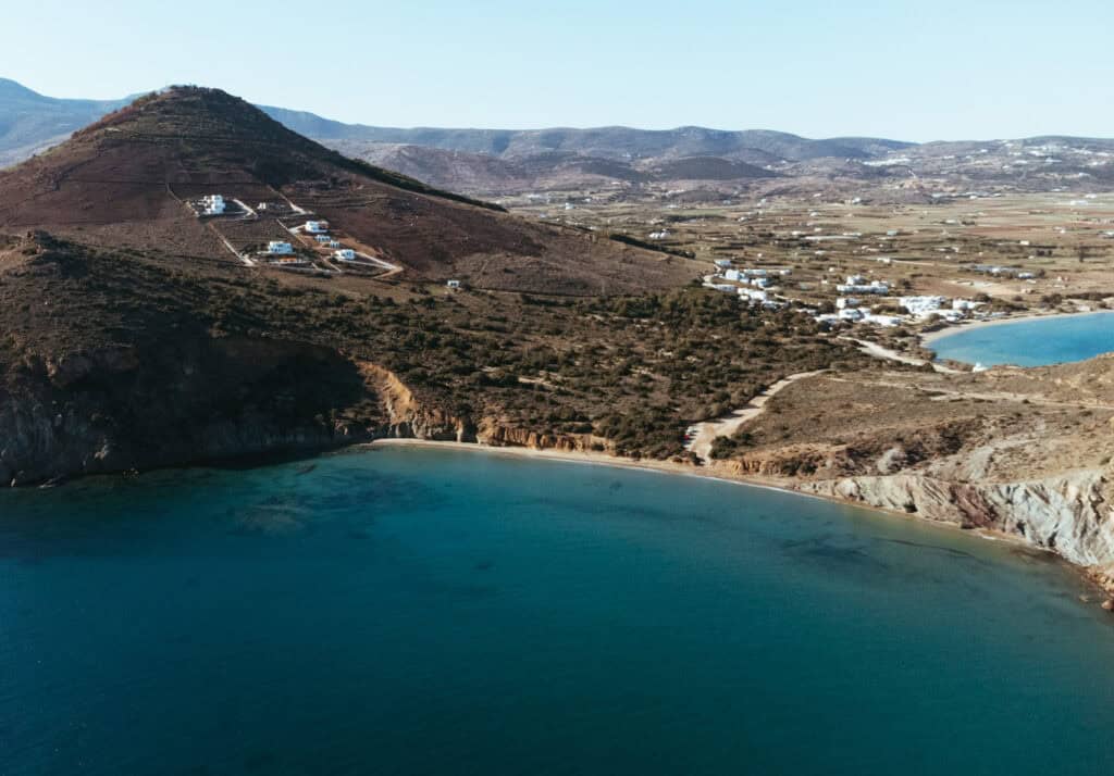 Aerial view of Kalogeros Beach in Paros, Greece, showcasing a secluded, curved sandy shoreline with clear blue water. The beach is bordered by rugged cliffs and a hillside dotted with a few white buildings. The surrounding landscape includes sparse greenery and a distant view of more buildings and another beach. The bright, clear sky enhances the tranquil and picturesque coastal scene.