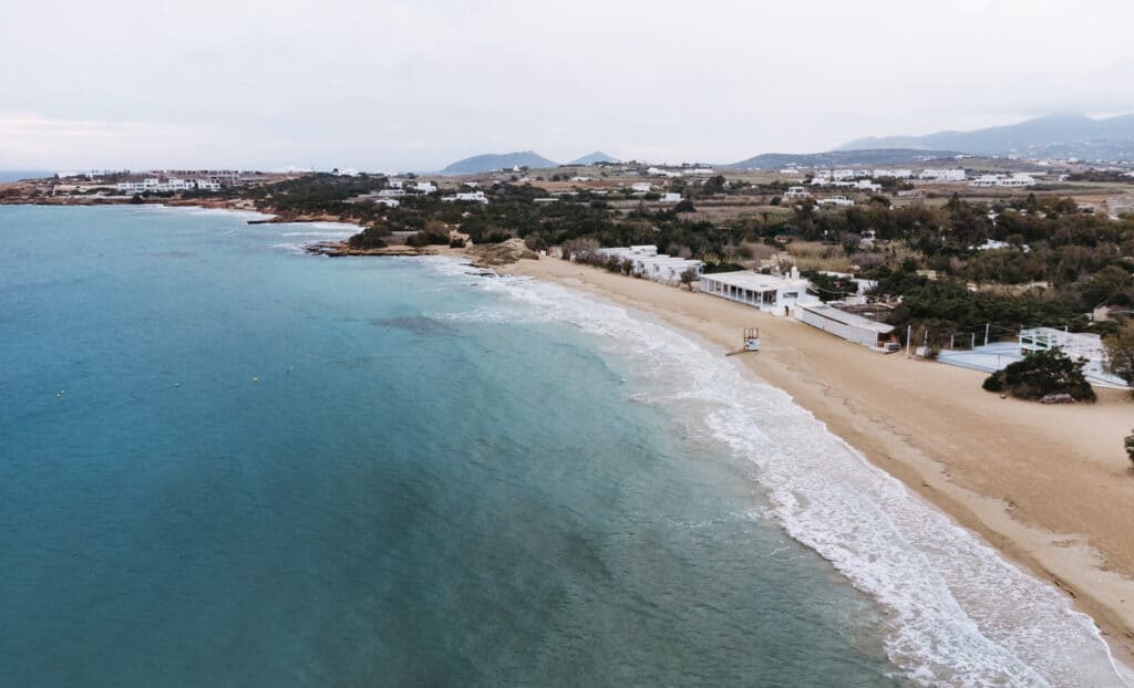 Aerial view of Santa Maria Beach in Paros, Greece, showcasing a long sandy beach with gentle waves lapping the shore. In the background, the landscape includes scattered white houses along the shore