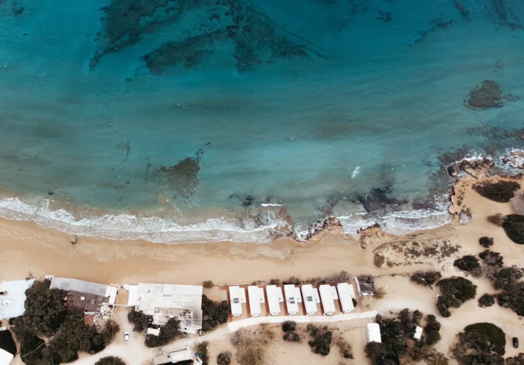 Aerial view of Santa Maria Beach in Paros, Greece, showing clear turquoise waters and sandy shores with gentle waves. The beach is lined with small, white beachfront and buildings. The scene includes some greenery and pathways near the shore.