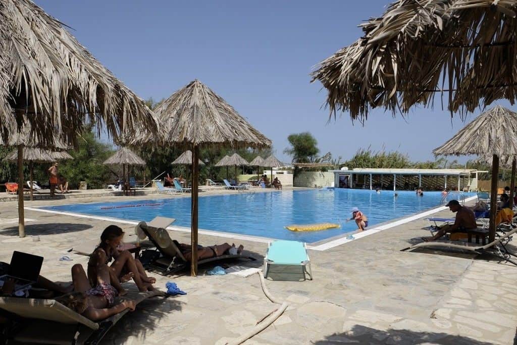 A lively outdoor pool area at Surfing Beach Villas shaded by thatched umbrellas and lined with sun loungers. Guests are seen relaxing by the pool and swimming, with a swim-up bar visible in the background.