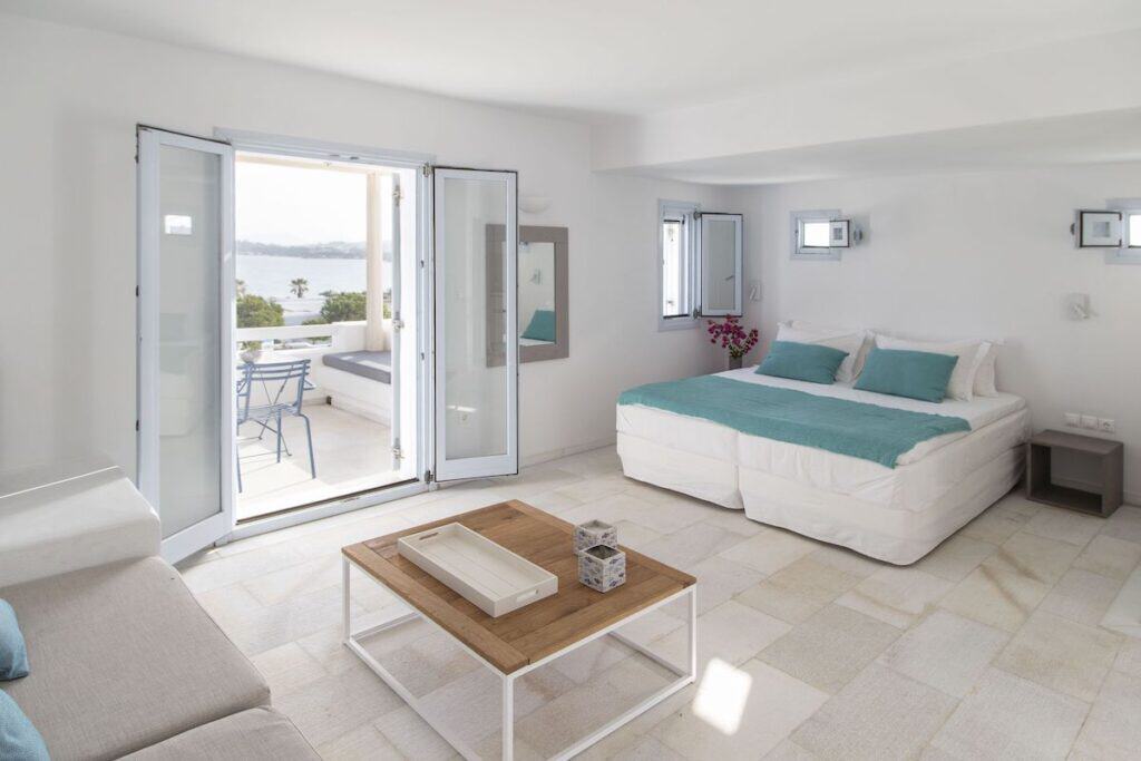 A bright and airy hotel suite at The Sand Collection with a minimalist Cycladic design. The room includes a spacious bed, a cozy seating area, and double doors opening to a private balcony with a sea view. 