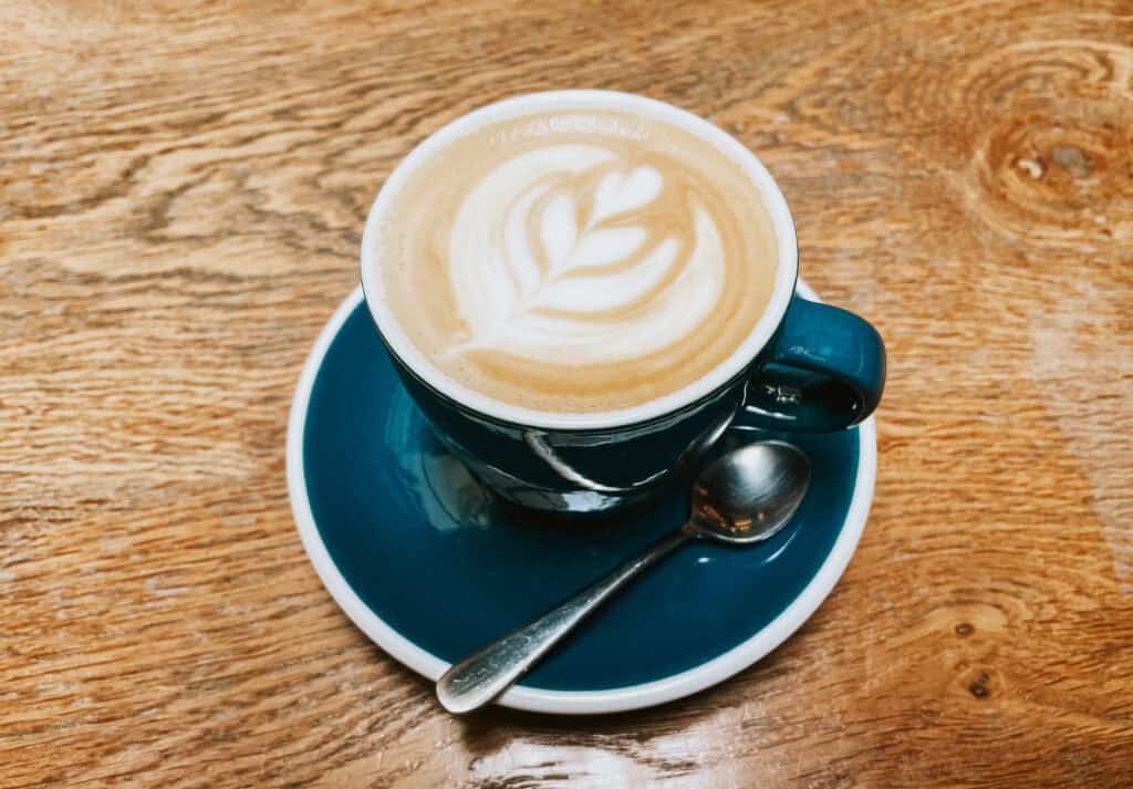 A close-up of a latte in a dark teal cup and saucer, placed on a wooden table. The latte features intricate latte art in the shape of a leaf, and a small silver spoon rests on the saucer.
