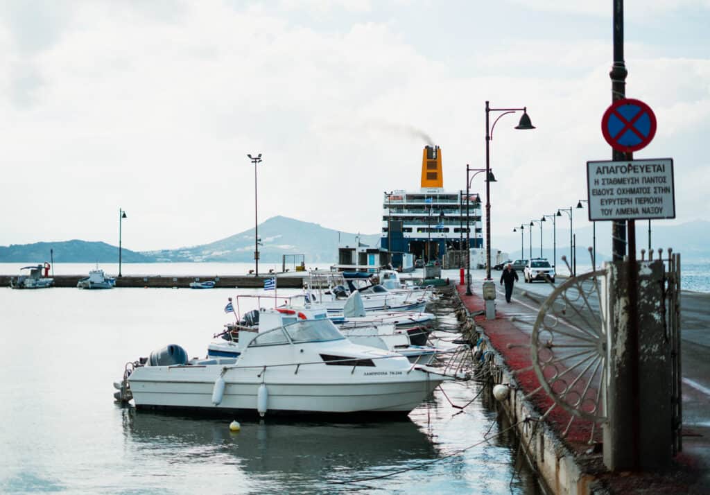 Small boats docked along a pier with a large ferry in the background, emitting smoke from its funnel, under a partly cloudy sky with distant hills visible. A sign in Greek on the right side prohibits vehicle parking in the port area.