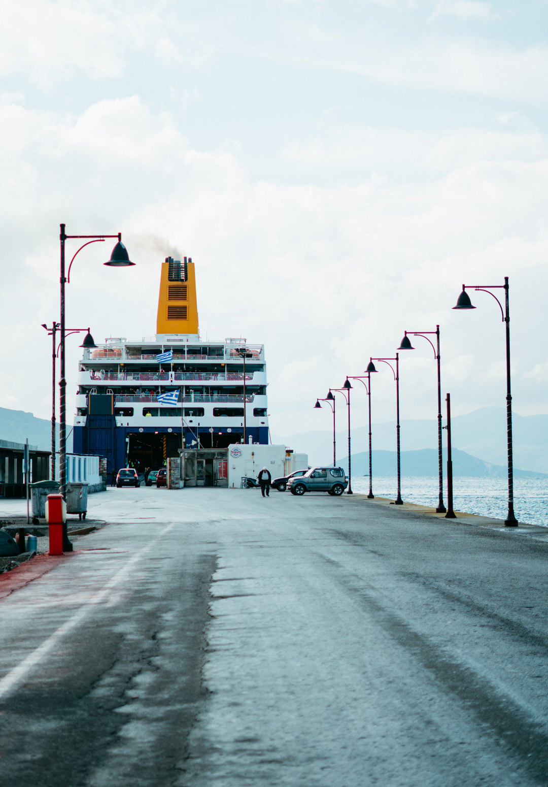 Large ferry docked at a port with cars and a few people on the pier, under a partly cloudy sky with distant hills visible. The ferry is emitting smoke from its funnel and has a Greek flag on its deck.