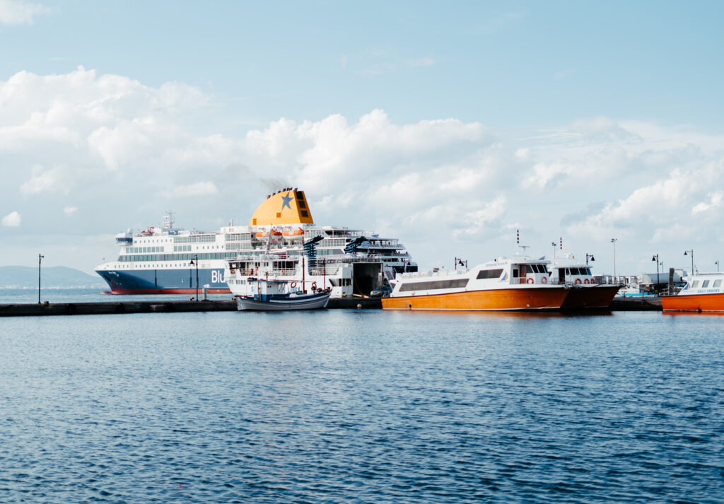 Several ferries docked at a port under a partly cloudy sky, including a large ferry with a yellow funnel and blue star, labeled 'Blue Star Ferries,' alongside smaller orange and white boats.