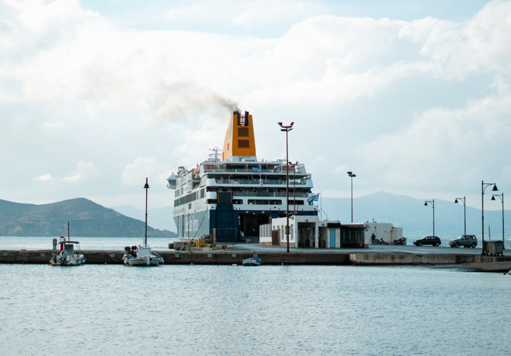 Large ferry docked at a port with a yellow funnel emitting smoke, under a partly cloudy sky with hills visible in the background.