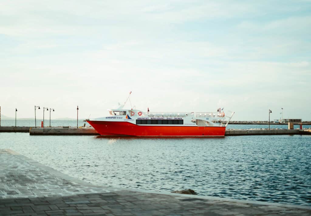 Red and white ferry named 'KYRIARHOS V' docked at a pier, with signage indicating 'Naxos Cruises' and 'Daily Cruises' under a clear sky.