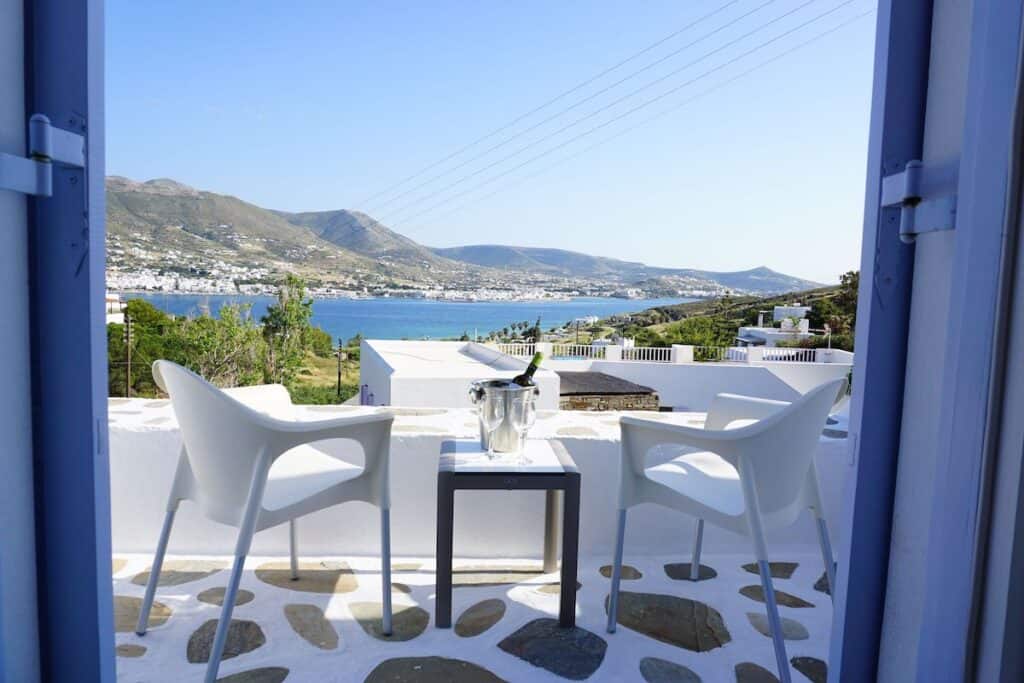 A scenic balcony view from Akrotiri Hotel overlooking the deep blue sea and rolling green hills of Paros, Greece. Two white chairs and a small table with a chilled bottle of wine and two glasses. The stone-accented balcony floor and traditional white-washed architecture enhance the Mediterranean charm, framed by soft blue door shutters.