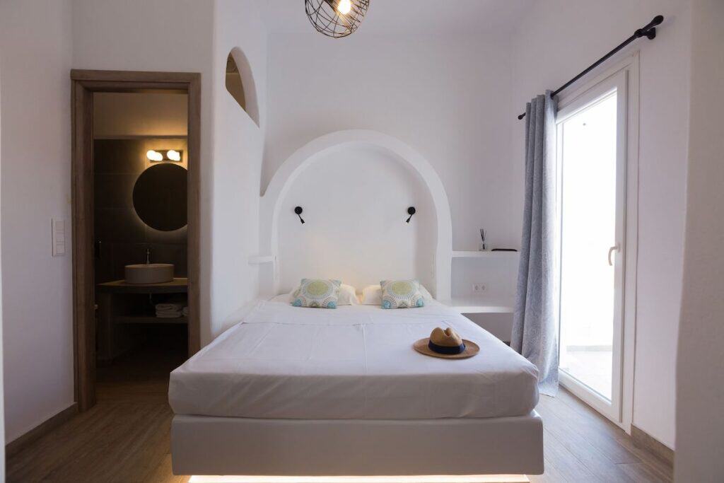 A cozy, minimalist bedroom with a white-washed arched headboard, built-in shelves, and a soft-lit ambiance. A straw hat rests on the neatly made bed with decorative pillows. A sliding glass door leads to a private balcony, allowing natural light to flood the space.