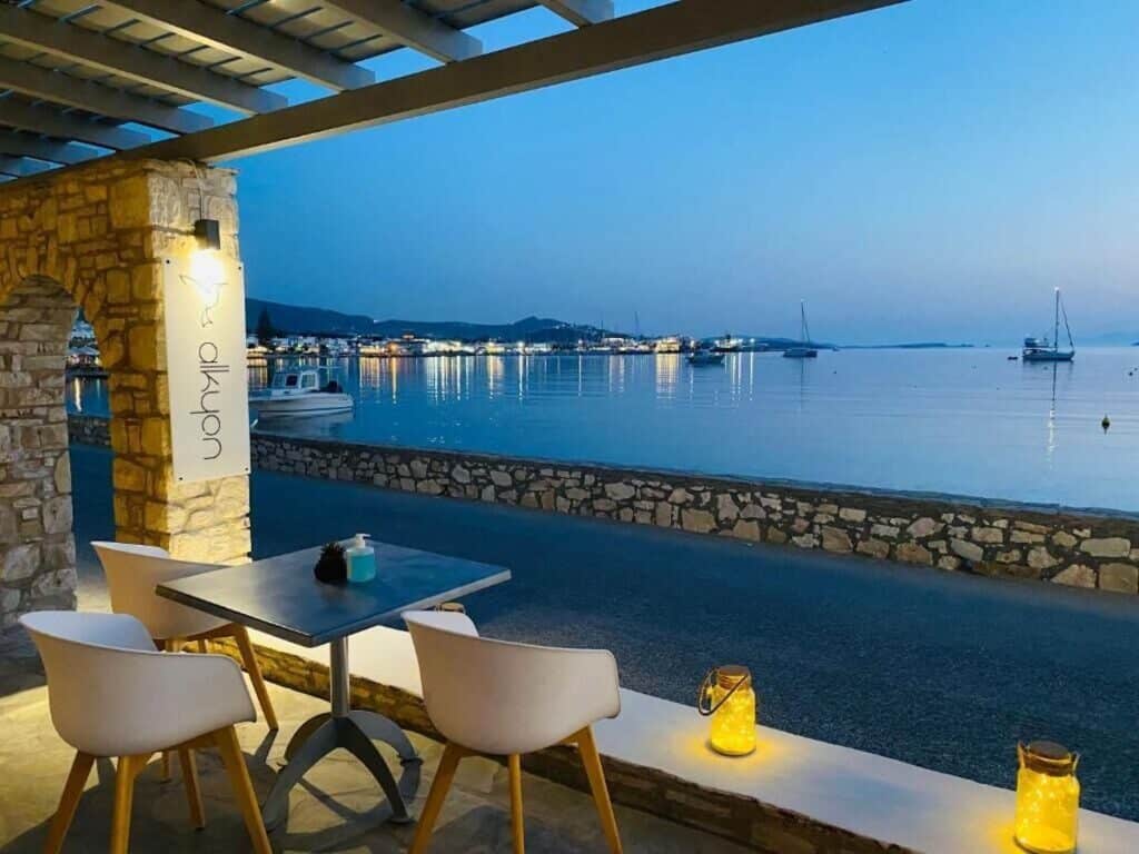 A waterfront terrace at Alkyon Hotel with a stunning evening view of the calm sea and boats. Stone archways, candle-lit tables, and soft blue lighting create a peaceful dining experience by the water.