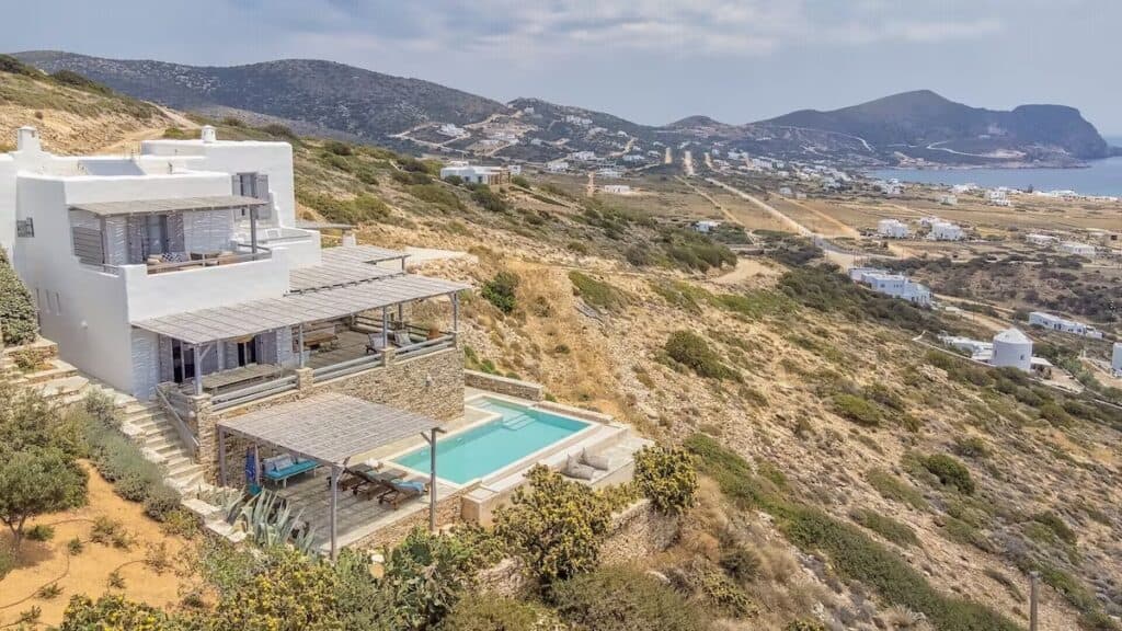 A luxurious white villa perched on a hillside overlooking the sea, featuring a private pool, shaded terraces, and stunning views of the surrounding landscape with rolling hills and scattered white houses.