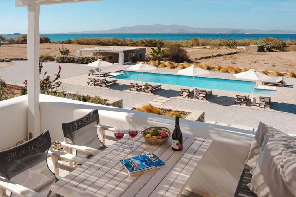 A stunning balcony view overlooking a serene pool area and the deep blue sea. A wooden table with a bottle of wine, two glasses, and a bowl of fresh fruit sits under a shaded pergola. Lounge chairs and umbrellas surround the pool, creating a luxurious Mediterranean retreat.