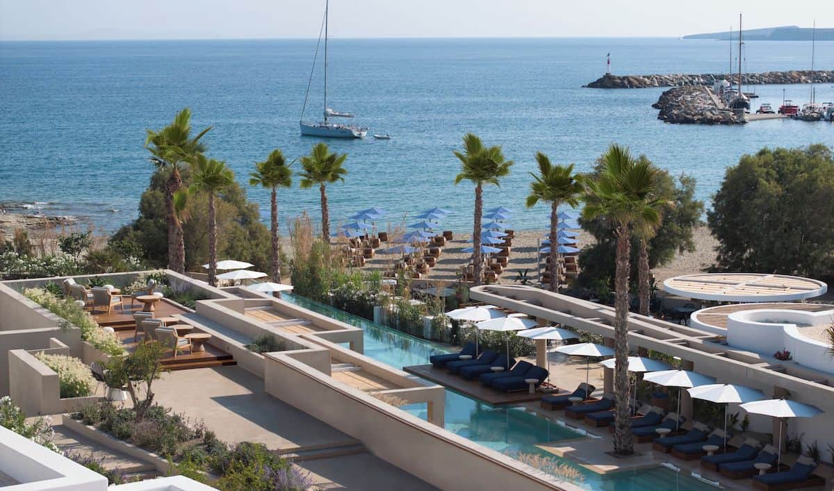 A luxurious beachfront resort with an elegant infinity pool, palm trees, and sun loungers. The property overlooks a sandy beach with rows of umbrellas and a calm, turquoise sea dotted with sailboats.