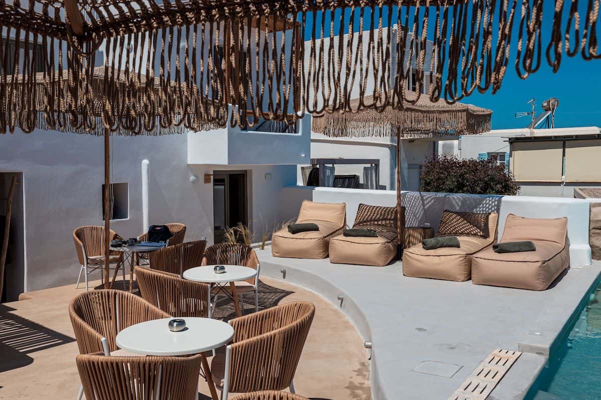A stylish outdoor lounge area featuring woven rope sunshades, wicker chairs, and beige bean bag loungers around a serene pool. The Cycladic-style white buildings add to the hotel's chic and relaxed aesthetic.