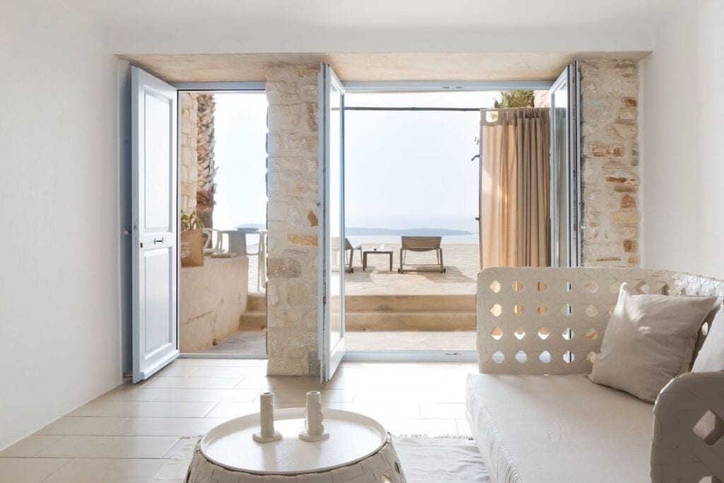 A tranquil, minimalist indoor seating area with a white sofa, stone walls, and an open doorway leading to a private terrace. Sun loungers outside offer a spectacular sea view under soft, golden light.
