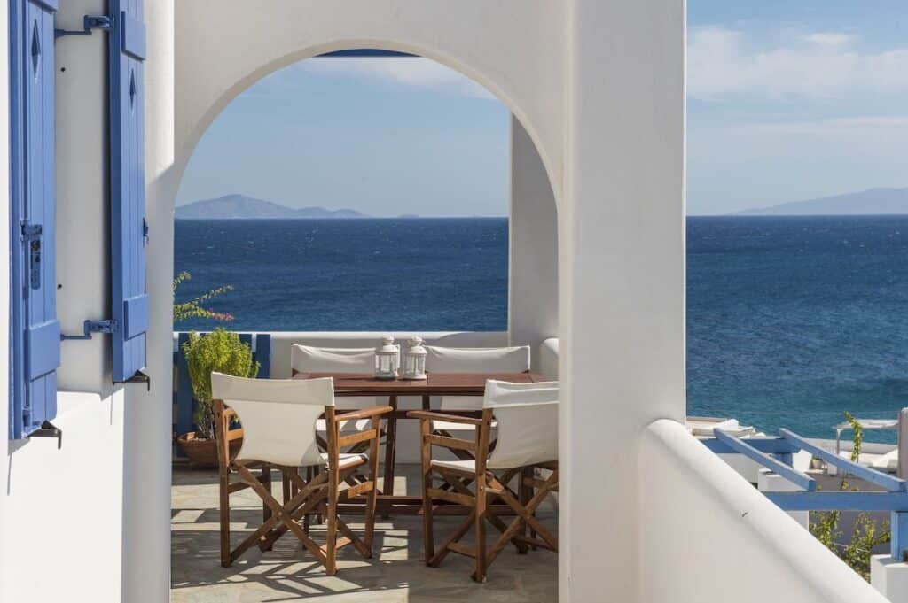 A picturesque terrace at Cleopatra Seaside Homes with white-washed walls and blue shutters, featuring a wooden table and chairs overlooking the deep blue Aegean Sea