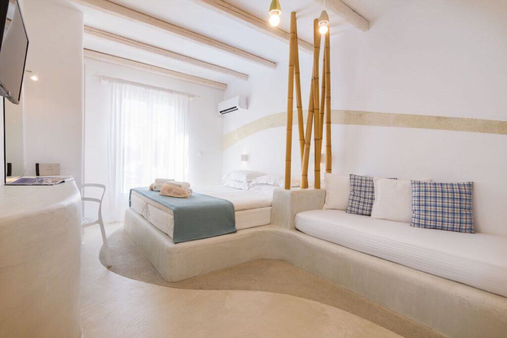 A bright and airy hotel room with Cycladic design, featuring a built-in bed with blue accents and a raised seating area with plaid pillows. Bamboo poles add a natural element to the partition wall. Sunlight pours in through sheer curtains, enhancing the tranquil atmosphere.