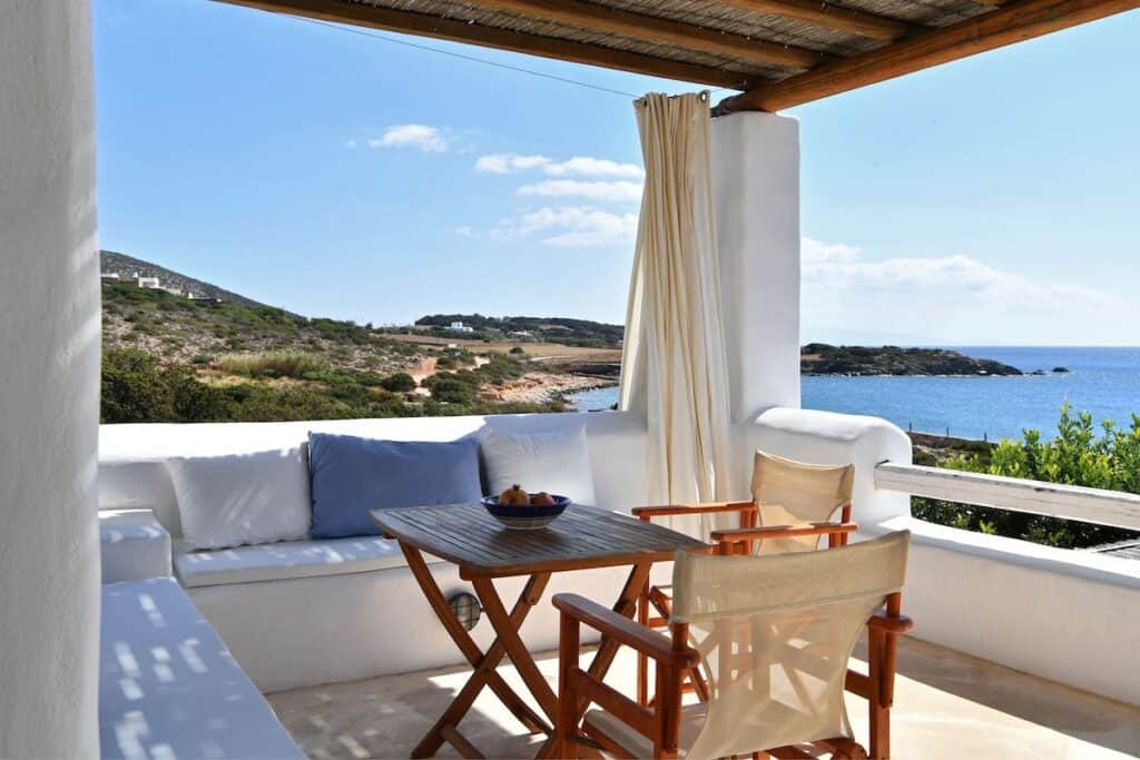 A charming outdoor terrace of a Greek-style villa, with a wooden dining table and director’s chairs, shaded by a pergola, offering breathtaking views of the coastline and deep blue sea.