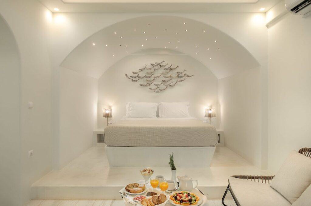 A luxurious bedroom with an arched ceiling, featuring delicate star-like lights and a wall sculpture of fish. A breakfast tray with fresh fruits, pastries, and juice sits at the foot of the bed. The soft, neutral color palette creates a peaceful and elegant ambiance.