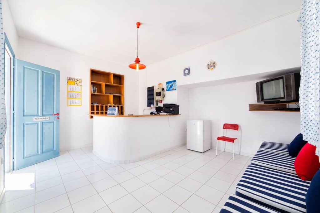 A bright and minimalist hotel reception area at Fivos Apartments with white walls and tiled flooring. A wooden front desk, blue entrance door, and striped lounge seating give the space a simple yet welcoming atmosphere.