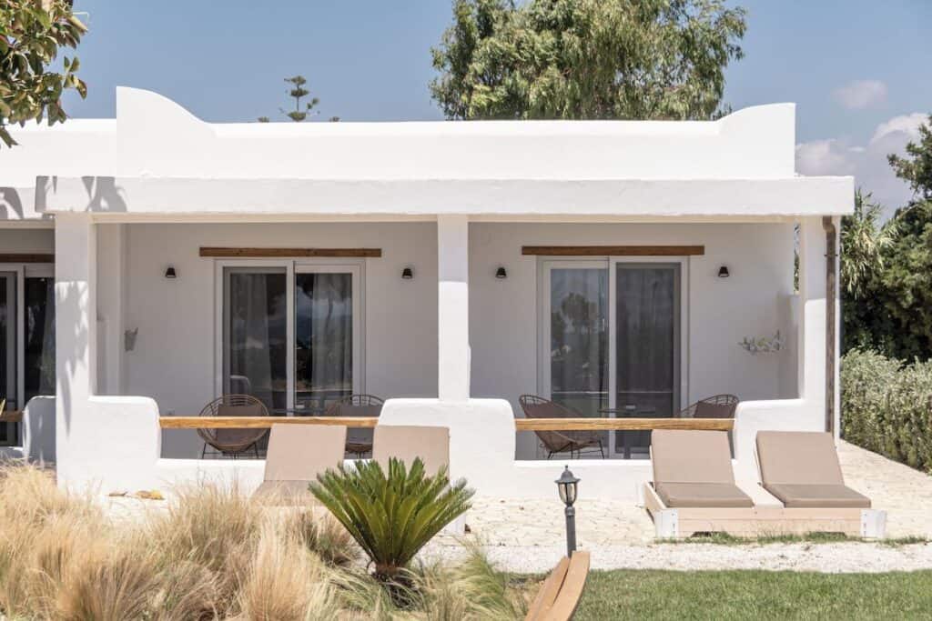 A charming beachfront bungalow with a white stucco exterior, wooden accents, and a private patio. Two sun loungers face a lush garden with tall grasses and tropical plants. Large sliding doors lead inside, where a cozy interior awaits.