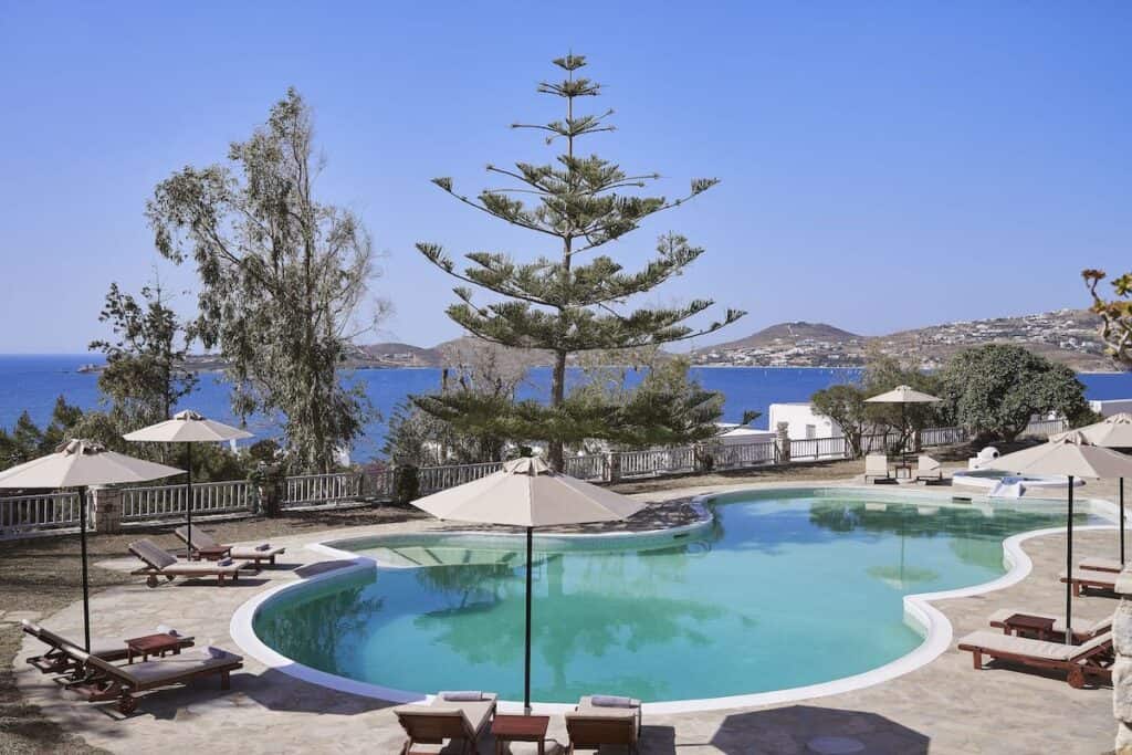 A scenic outdoor pool area with a curved swimming pool, surrounded by sun loungers and umbrellas. Lush trees frame the stunning view of the blue Aegean Sea and distant hills.
