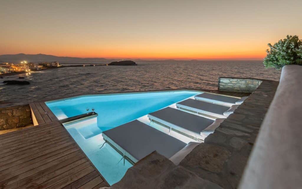 A stunning infinity pool overlooking the sea at sunset, with built-in submerged loungers for a luxurious, relaxing experience. The pool’s soft blue glow contrasts beautifully with the warm hues of the horizon. In the distance, city lights twinkle along the coastline.