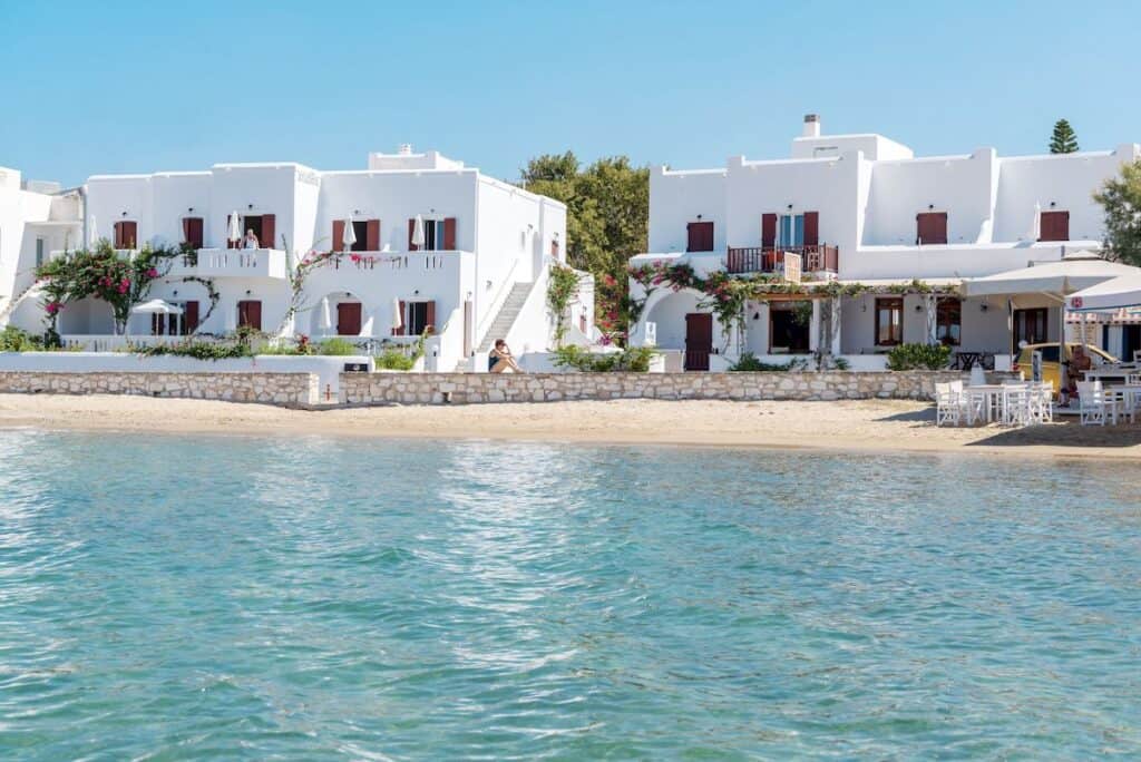 A charming seaside hotel with classic Cycladic white buildings adorned with blooming pink bougainvillea. The hotel sits directly on the sandy shore, offering a peaceful beachfront escape